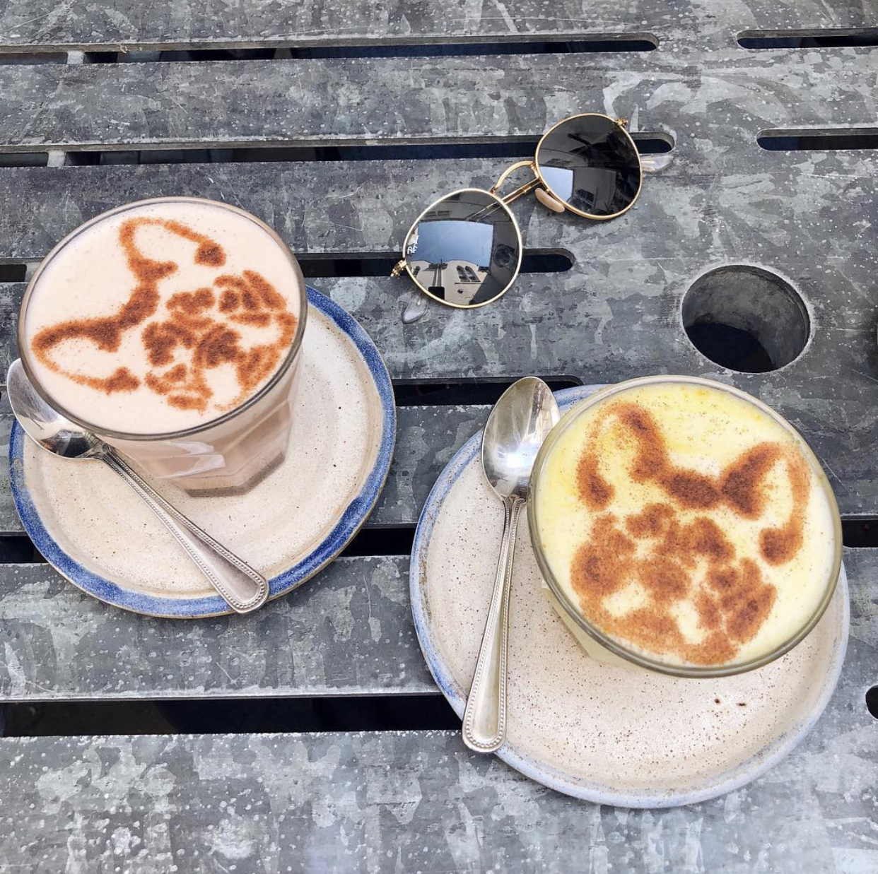 Two coffees with a dog design on the top, on a table next to a pair of Ray-Ban sunglasses