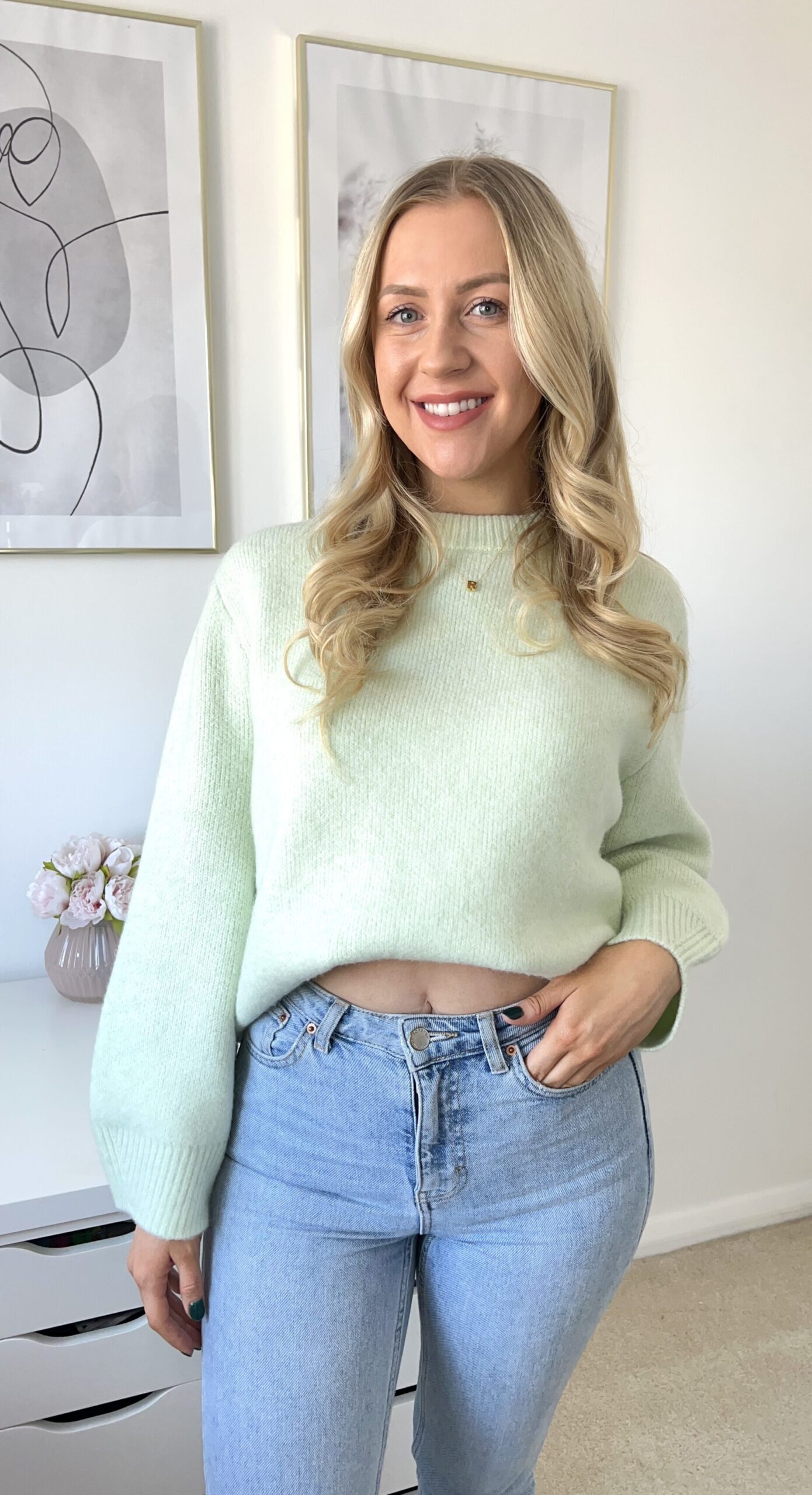 Rosie Eva Millard, a white woman with blonde hair, smiling at the camera. She is wearing a thick mint green jumper and blue denim jeans, one hand in a pocket